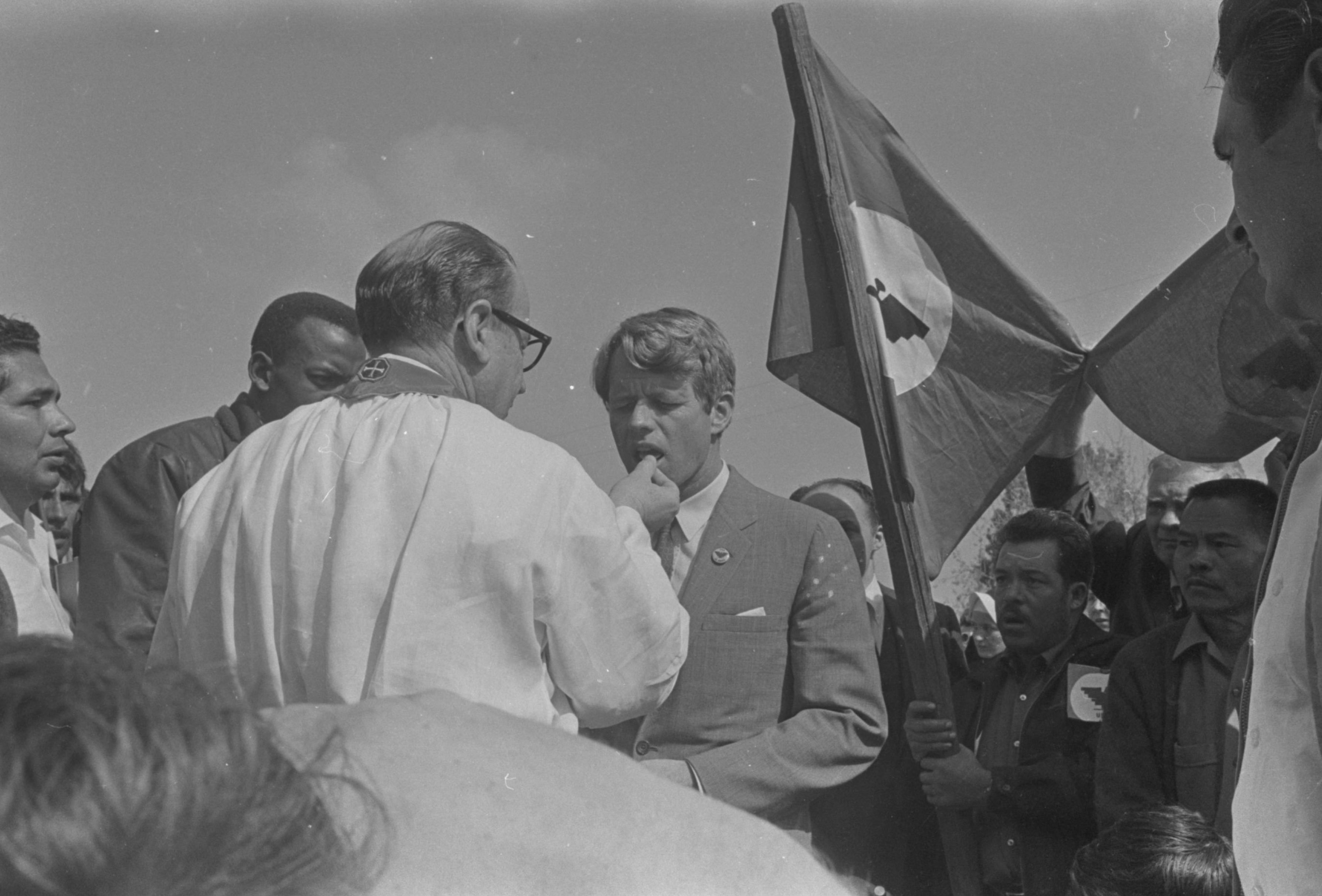 Father Eugene Boyle offers the Catholic sacrament of the holy communion to César Chávez on the day Chávez broke his twenty five day fast. A debilitated Chavez is helped by people surrounding him. A UFWOC flag waves behind Chávez. El padre Eugene Boyle ofrece el sacramento católico de la sagrada comunión a César Chávez el día que Chávez rompió su ayuno de veinticinco días. Un Chávez debilitado necesita la ayuda de los que le rodean para mantenerse en pie. Una bandera de la UFWOC ondea detrás de Chávez.