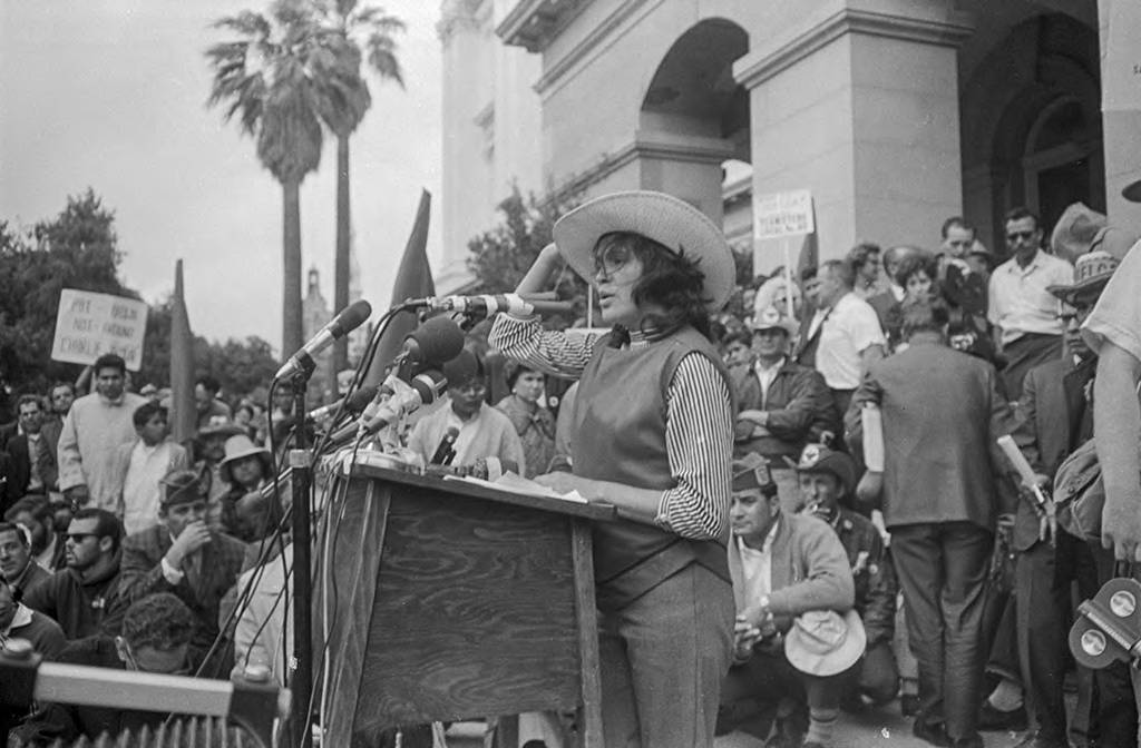 Dolores Huerta, vice president of the National Farm Workers Association (NFWA) and principal speaker of the rally, speaks to a crowd in front of State Capitol on the last day of the march from Delano to Sacramento. California Governor Pat Brown refused to meet with the marchers that day and instead spent the weekend in Palm Springs. “We are no longer interested in listening to the excuses the Governor has given in defense of the growers, to his apologies to them for not paying us decent wages or why the growers cannot dignify the workers as individuals with the right to place the price on their own labor through collective bargaining,” said Huerta according to the New York Times.Dolores Huerta, vicepresidente de la Asociación Nacional de Campesinos (NFWA) y oradora principal de la manifestación, habla ante una multitud frente al capitolio estatal el último día de la marcha de Delano a Sacramento. El gobernador de California, Pat Brown, se negó a reunirse con los manifestantes ese día y, en cambio, pasó el fin de semana en Palm Springs. “Ya no nos interesa escuchar las excusas que ha dado el Gobernador en defensa de los dueños, sus disculpas por no pagarnos salarios dignos o por qué los dueños no pueden dignificar a los trabajadores como individuos con derecho a poner el precio en su propio trabajo a través de la negociación colectiva”, dijo Huerta según el New York Times.