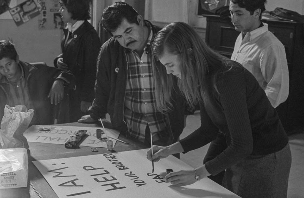 UFW volunteer and organizer Wendy Goepel is working on marking picket signs in the basement of the Church of the Epiphany in Lincoln Heights, in Los Angeles. "This was in Los Angeles," she said in an email after seeing this photograph. "After the harvest we went to LA to do informational picketing during shift changes at big manufacturing plants. Every car leaving the parking lot got leafletted; some gave small money; many gave encouragement; none could buy grapes after meeting us! There were about ten of us. I loved making signs and did it a lot in the church basement where we were living on their floor."</p>
<p>Wendy Goepel, voluntaria y organizadora de UFW, está trabajando en hacer pancartas en el sótano de la Iglesia de la Epifanía en Lincoln Heights, in Los Angeles. "Esto fue en Los Ángeles. Después de la cosecha, fuimos a Los Ángeles para hacer piquetes informativos durante los cambios de turno en las grandes plantas de fabricación," dijo Goepel en un correo electrónico después de ver esta foto. "Cada automóvil que salía del estacionamiento recibió folletos; algunos dieron dinero pequeño; muchos dieron ánimos; ¡ninguno pudo comprar uvas después de conocernos! Éramos unos diez. ¡Vivíamos en la parte baja de la cadena alimenticia, seguro! Me encantaba hacer letreros y lo hice. mucho en el sótano de la iglesia donde vivíamos, en su piso."