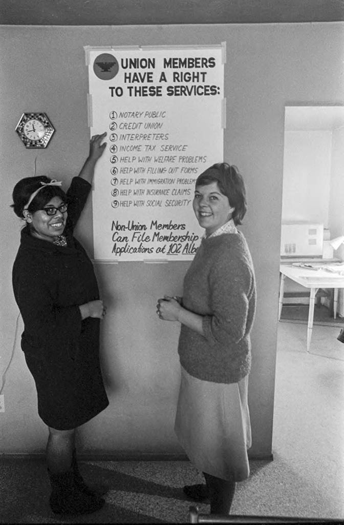 Jessica Govea and Bonnie Chatfield standing next to the NFWA's poster of the Service Center. The poster lists in English the nine services offered to the union's members who speak English: notary public, credit union, interpreters, income tax services, welfare issues, filling out forms, immigration issues, insurance claims, and social security.</p>
<p>Jessica Govea y Bonnie Chatfield de pie junto al cartel del Centro de Servicio de NFWA. El cartel enumera en inglés los nueve servicios que se ofrecen a los miembros de la unión que hablan inglés: notario publico, union de crédito, intérpretes, servicios de income tax, problemas con el welfare, llenado de documentos, problemas de migración, aseguranza y seguro social.