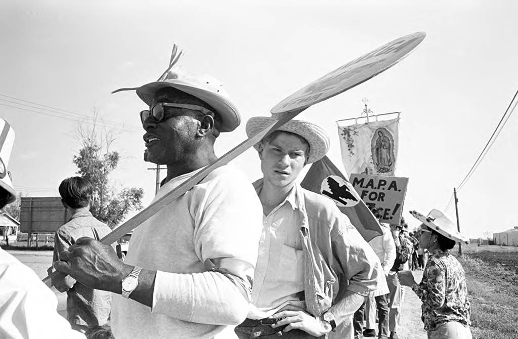 Wearing sunglasses and a hat, William King, the oldest original marcher, carries a Huelga-NFWA sign during the march to Sacramento in 1966. A Mexican American Political Association (MAPA) sign is visible in the back. Con gafas de sol y un sombrero, William King, el manifestante original de mayor edad, lleva un cartel de Huelga-NFWA durante la marcha a Sacramento en 1966. En la parte de atrás se ve un cartel de la Asociación Política Mexicoestadounidense (MAPA).