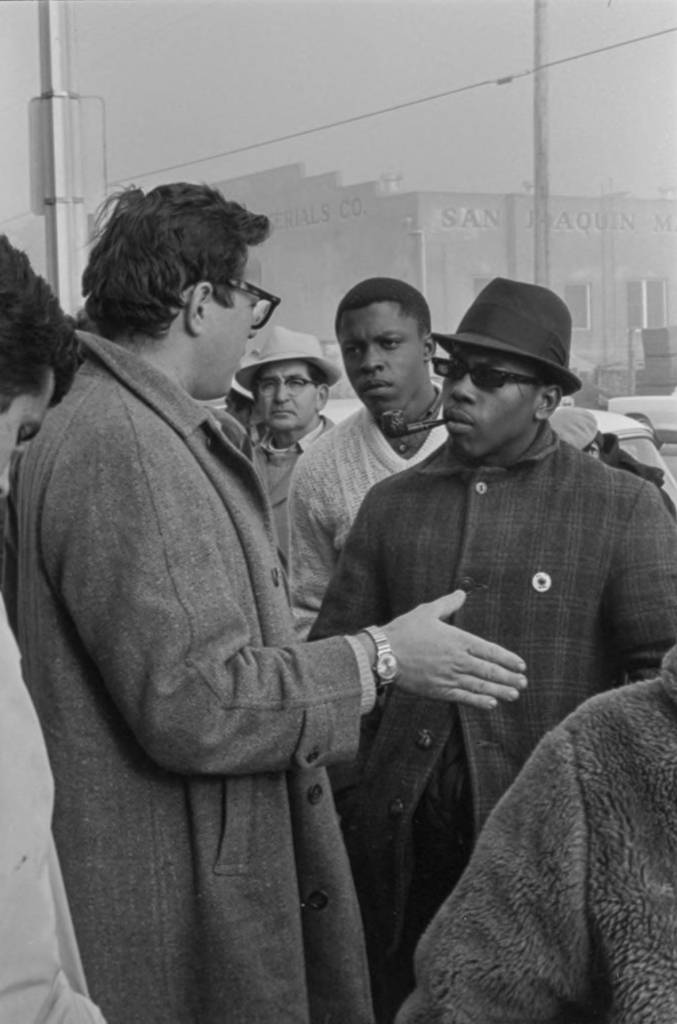 Rev. Eugene Boutilier is talking to a UFW supporter from the Black Power movement during a boycott. The ally is wearing the UFW pin with the black eagle, symbol of the farmworker movement, on his coat and smoking a pipe.	Rev. Eugene Boutilier está hablando con un partidario de UFW del movimiento Black Power durante un boicot. El partidario lleva el pin de UFW con el águila negra, símbolo del movimiento campesino, en su abrigo y fumando una pipa.