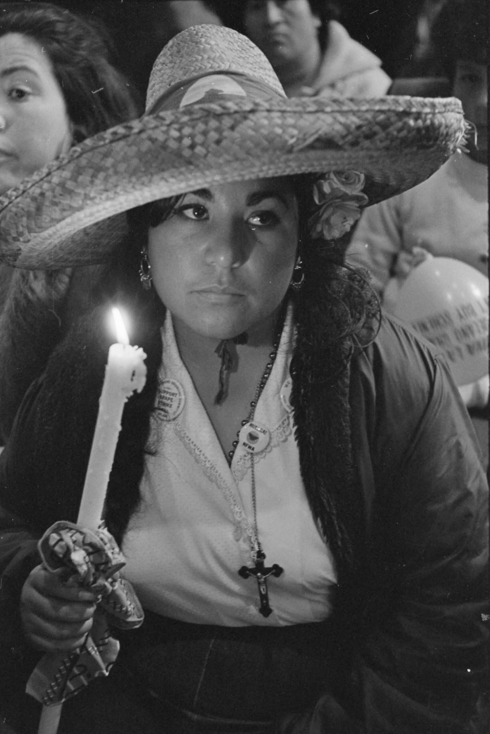 Carolina Franco holds a candle at a rally during the march to Sacramento. She is wearing a large hat, and a crucifix and rosary hang from her neck. Modesto, California, April 1, 1966. Photo by John Kouns.