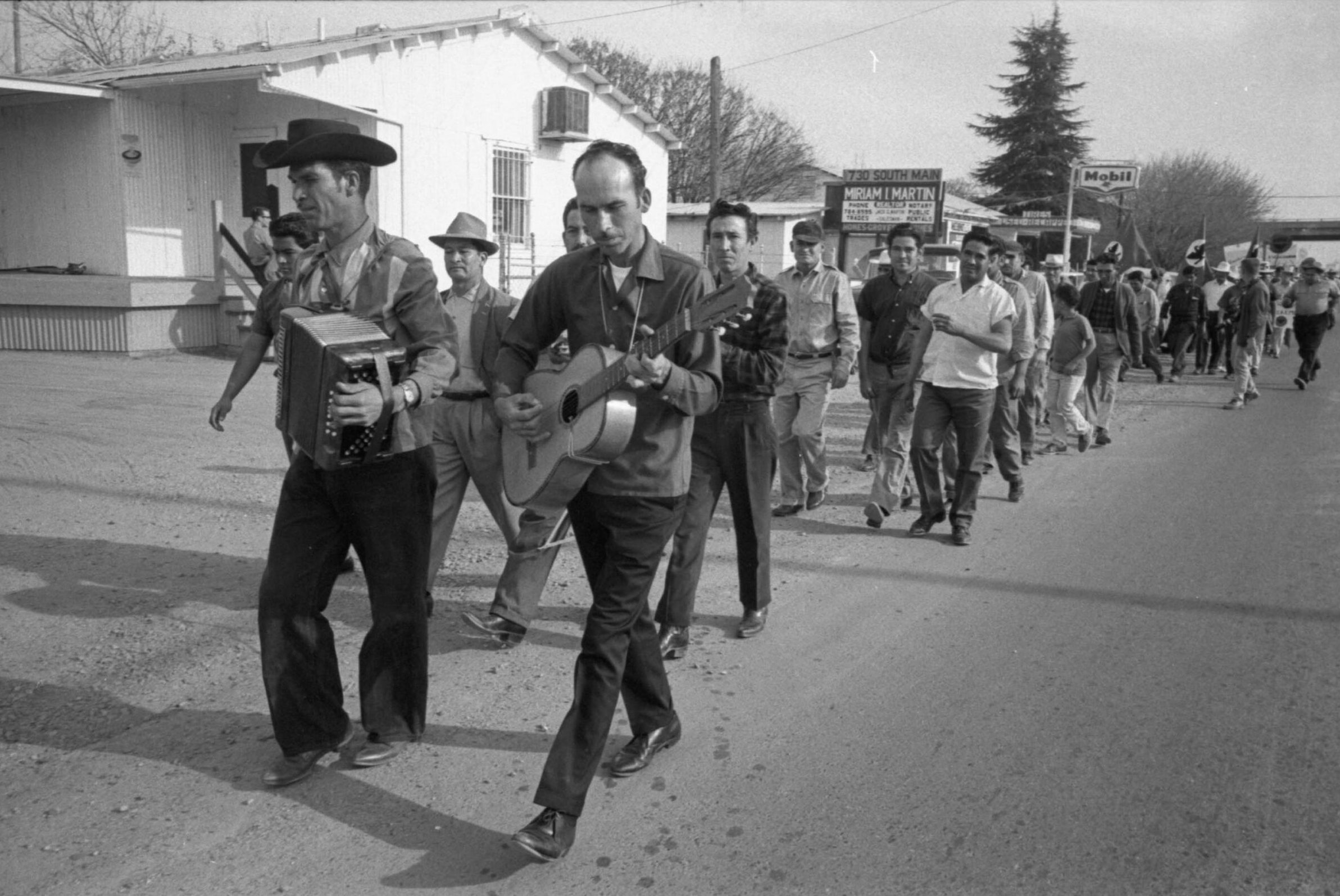 Jesús Marín "Marín" Barrera (playing the accordion), José Raúl "Rico" Barrera (playing the guitar), and Gilberto "Beto" Garza (playing the drum) with the marchers as they enter Porterville, California on 03/18/1966 during the march to Sacramento. Also in photo are: Desiderio Ramirez (in cap), Antonio Rosas (in white shirt), Sebastián “Tano” Zamora (in baseball cap), Lucio Chavera (middle), and César Chávez (at the end). The marchers are walking past 730 South Main St, Porterville, California.</p>
<p>Jesús Marín "Marín" Barrera (tocando el acordeón), José Raúl "Rico" Barrera (tocando la guitarra), y Gilberto "Beto" Garza (tocando el tambor) con los manifestantes al entrar a Porterville, California el 18 de marzo de 1966 durante la marcha a Sacramento. También en la foto están: Desiderio Ramírez (con gorra), Antonio Rosas (con camisa blanca), Sebastián “Tano” Zamora (con gorra de béisbol), Lucio Chavera (en medio) y César Chávez (al final). Los manifestantes pasan por 730 South Main St, Porterville, California.