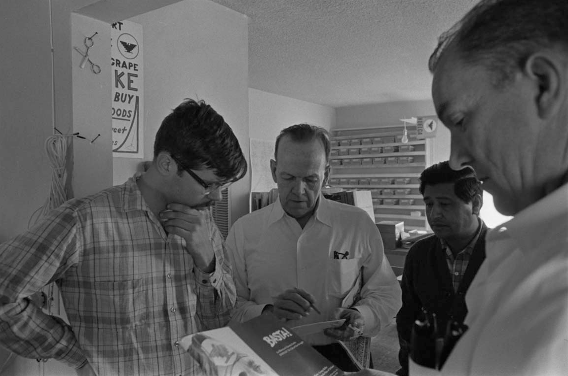 Doug Adair, an unidentified man, César Chávez, and Jack T. Conway are standing and talking in the office of El Malcriado. Conway was executive director of the AFL-CIO’s Industrial Union Department. Delano, ca. 1966. Doug Adair, un hombre no identificado, César Chávez y Jack T. Conway, platican en la oficina de El Malcriado. Conway era director ejecutivo del Departamento de Sindicato Industrial de la AFL-CIO.