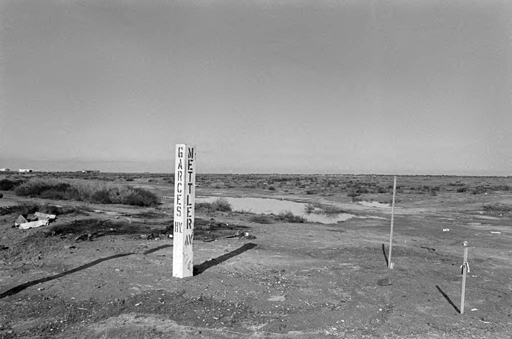 A view of the intersection of Garces Highway and Mettler Avenue in the outskirts of Delano. A pole can be seen with the names of the streets painted on it. In the background, dirt, bushes, and puddles of water can be seen in the desolate landscape. Farther west, Forty Acres was located.Una vista de la intersección de la carretera Garcés y la avenida Mettler en las afueras de Delano. Un poste se observa con los nombres de las calles pintados sobre él. Al fondo de la imagen en el paisaje solitario hay tierra, arbustos y charcos de agua. Más al oeste de la intersección se ubicaría el campus Forty Acres.