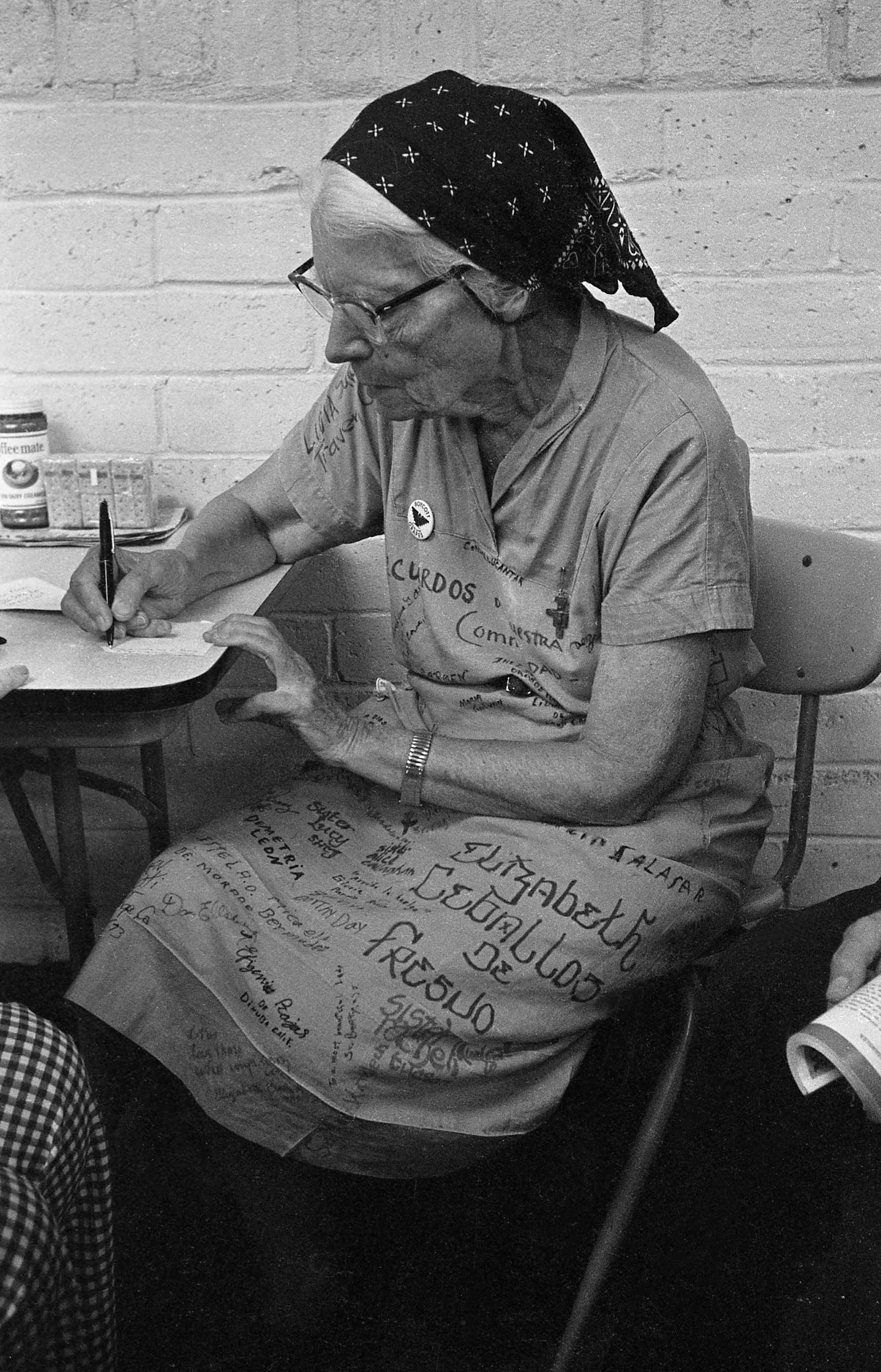 Dorothy Day writes on a piece of paper at the Fresno County Jail. Her jail uniform is signed by the women imprisoned with her. August 1973. Dorothy Day escribe en una hoja de papel en la carcel de condado de Fresno. Su uniforme de la cárcel está firmado por sus compañeras arrestadas, agosto de 1973.