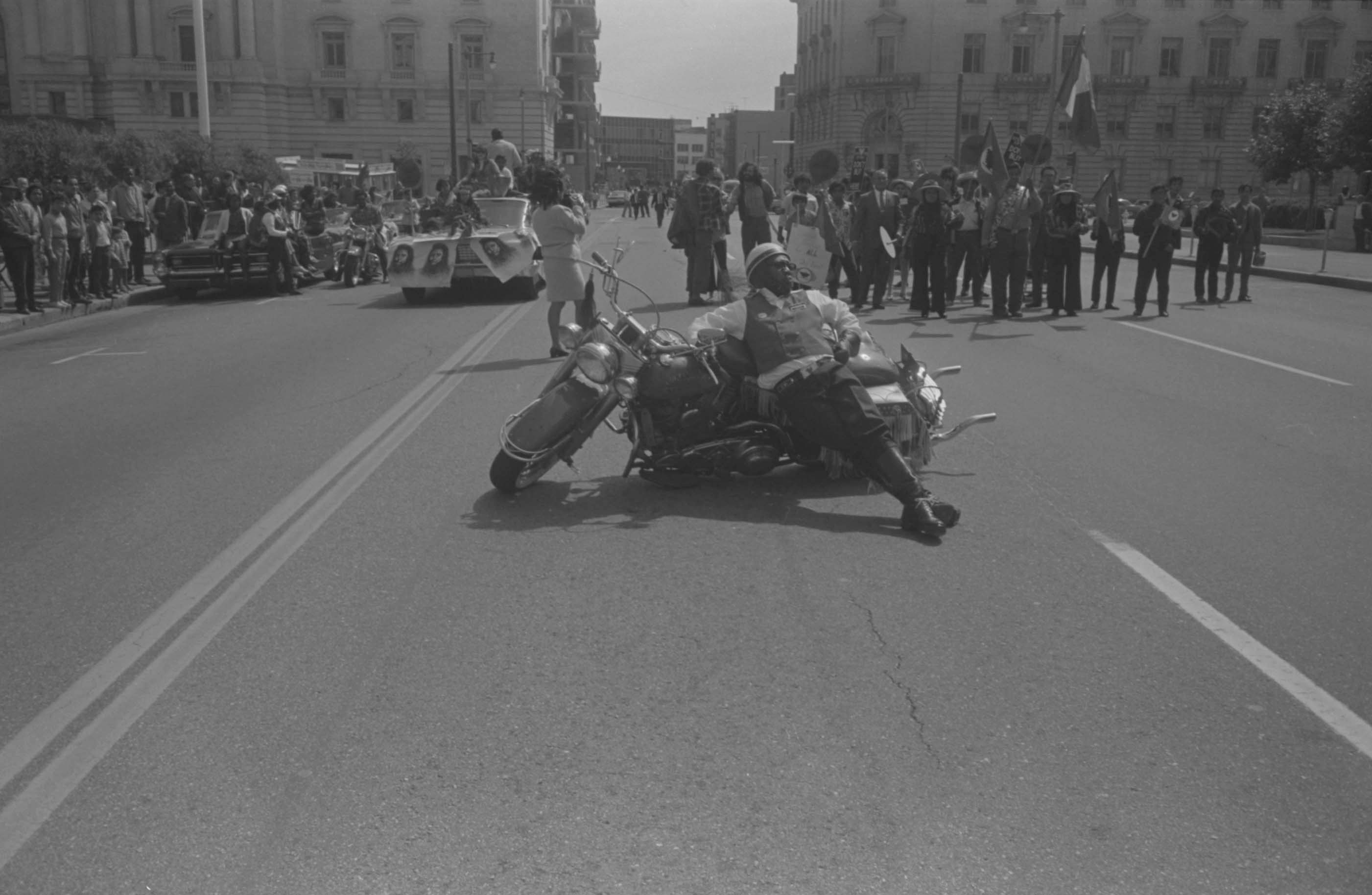 A member of the Space Riders Drill Team leans on a motorcycle on the road next to<br />
UFWOC marchers. They are near City Hall, where there is a rally for the Delano<br />
Support Committee. San Francisco, September 15, 1968. Photo by John Kouns.