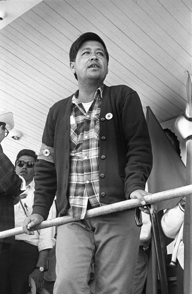 César Chávez leans on a rail during the march to Sacramento in 1966. Roberto Bustos (wearing sunglasses) is standing in the background. César Chávez está apoyado en una barandilla durante la marcha a Sacramento en 1966. Roberto Bustos (con lentes de sol) está de pie en el fondo de la imagen.