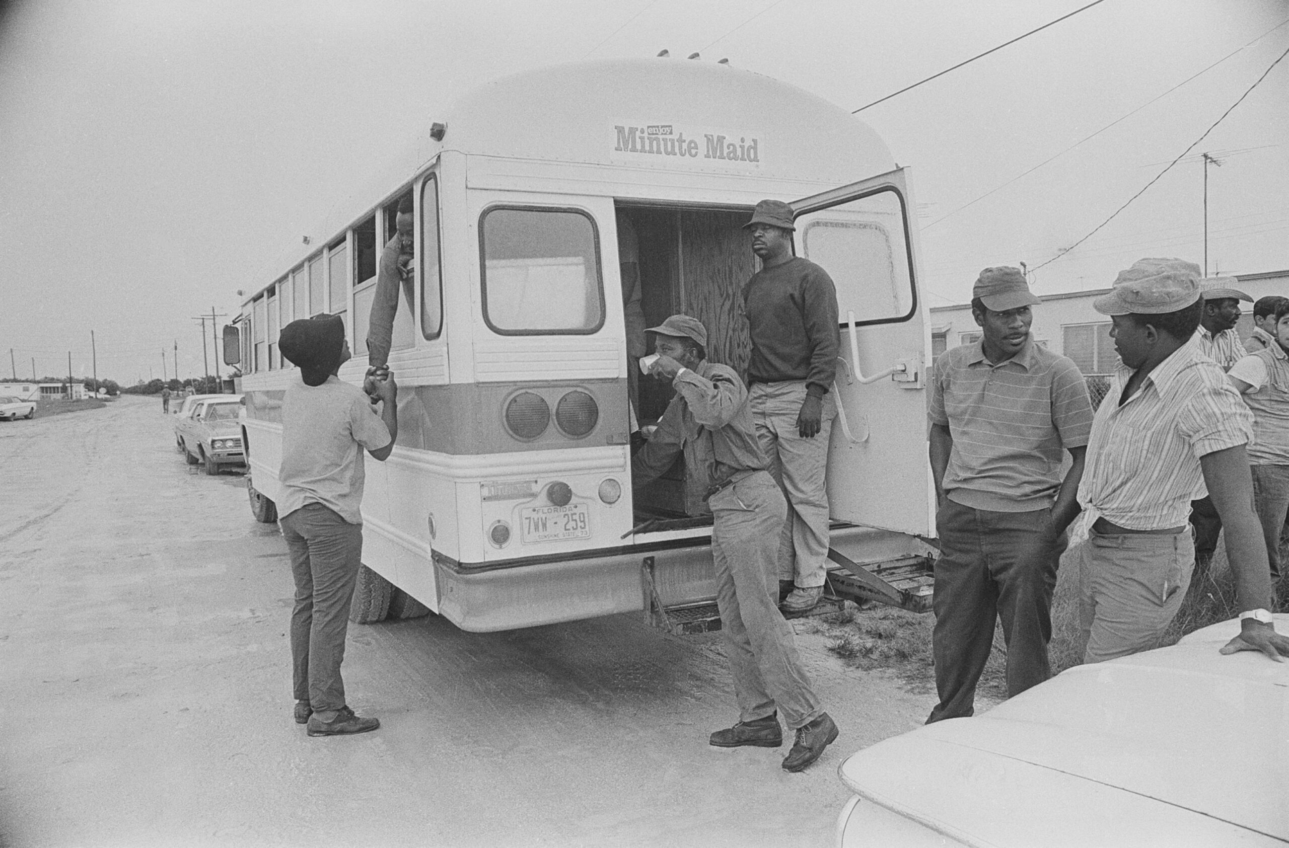 A group of farm workers stands next to a Minute Maid bus. Un grupo de trabajadores agrícolas se encuentra junto a un autobús de Minute Maid.