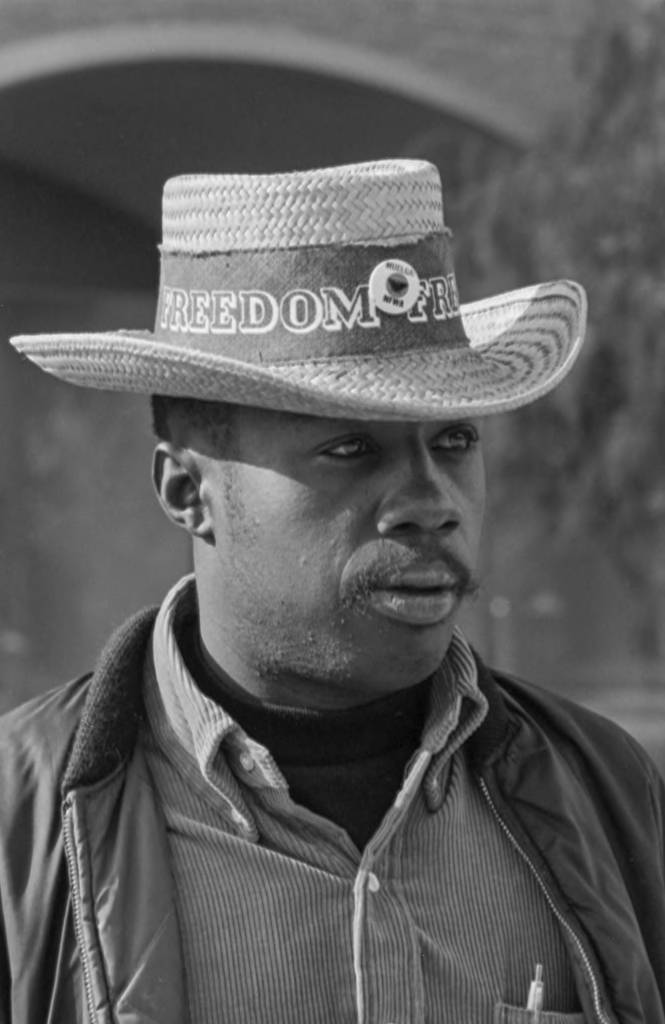 A UFW supporter is standing next to a store where picketers are organizing a boycott. He is wearing a straw hat. On the hat there is a writing that says, "Freedom" and the typical farmworker movement's button with the symbolic black eagle and the words: Huelga NFWA. Un simpatizante de UFW está de pie junto a una tienda donde los piqueteros están organizando un boicot. Lleva un sombrero de paja. En el sombrero hay un escrito que dice: "Libertad" y el botón típico del movimiento campesino con la simbólica águila negra y la layenda: Huelga NFWA.