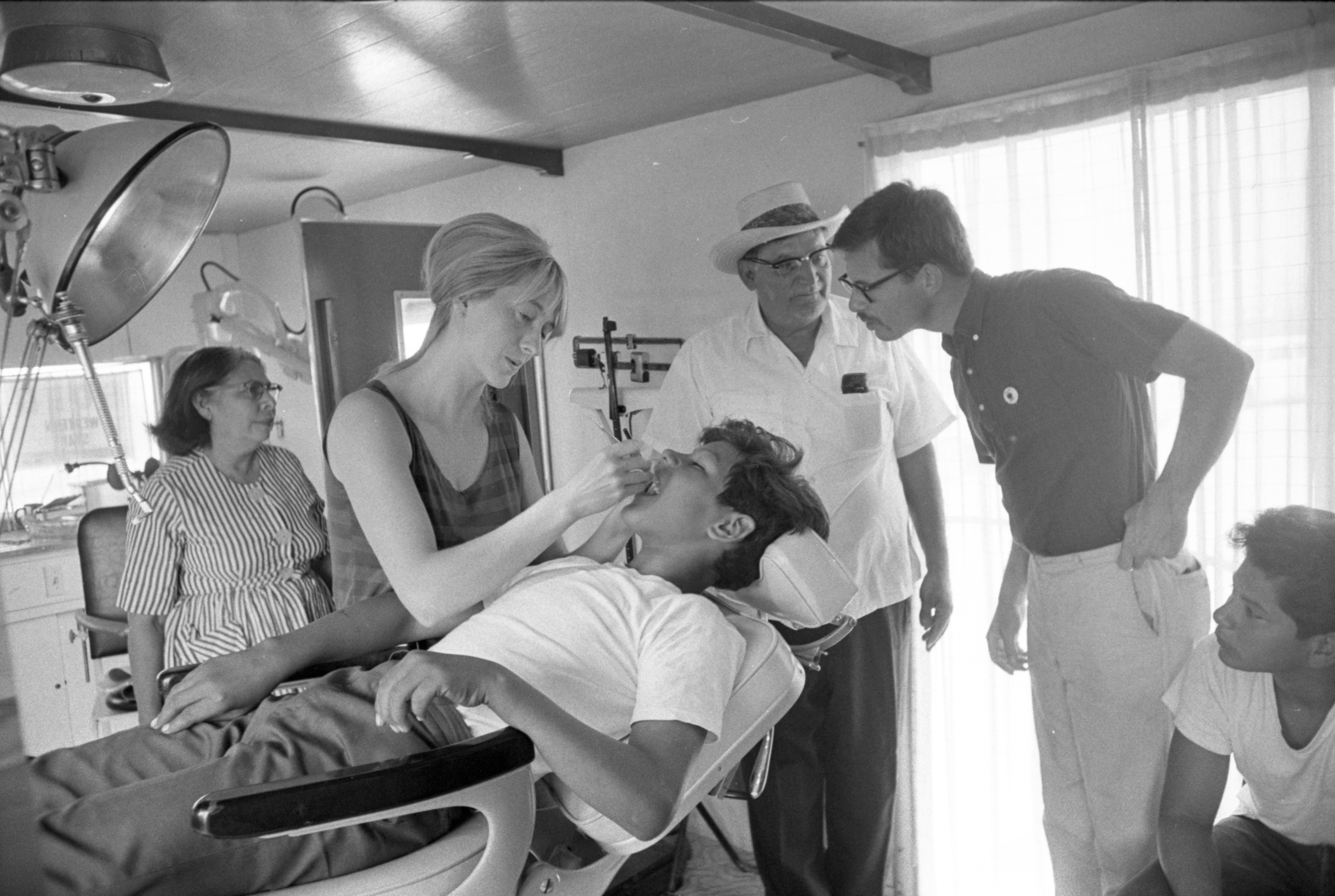 A nurse works on the teeth of a boy at the Farm Workers Health Clinic at Arroyo<br />Camp. Dr. David Brooks leans to observe the procedure while a woman, a man, and<br />a boy observe. Delano, California, 1967. Photo by John Kouns.