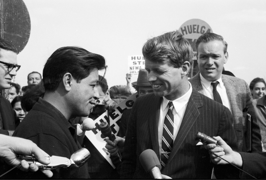 César Chávez (left) and Senator Robert F. Kennedy (D-NY, right) speak as they arrive at Delano High School ahead of the day's scheduled U.S. Senate Subcommittee on Migratory Labor hearing, which took place inside the auditorium. César Chávez (derecha) y el senador Robert F. Kennedy (D-NY, derecha) hablan mientras llegan a la preparatoria Delano antes de la audiencia programada del Subcomité del Senado estadounidense sobre el trabajo migratorio, la cual fue convocada dentro del auditorio.